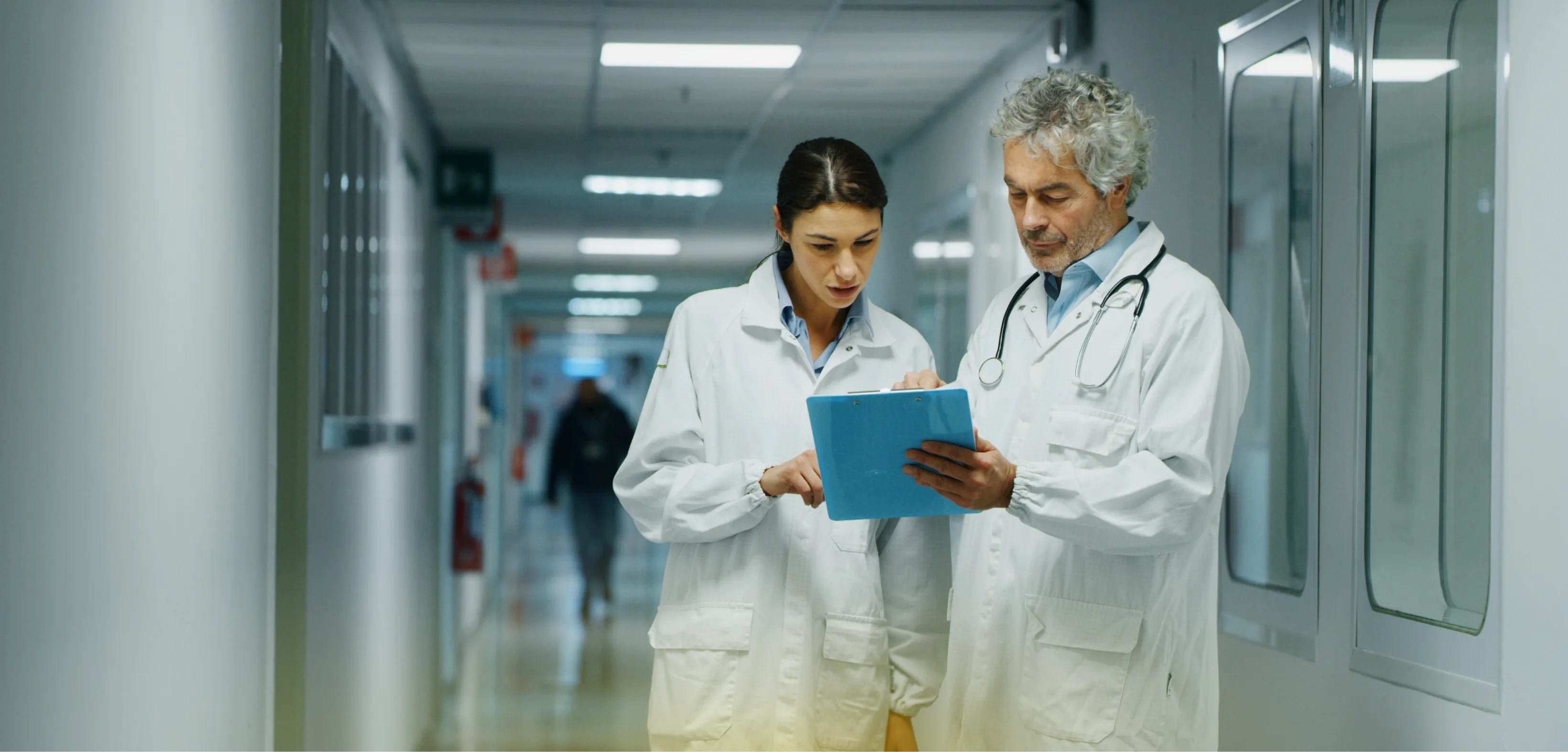 Two clinicians reviewing a patient chart on a clipboard in a hospital corridor